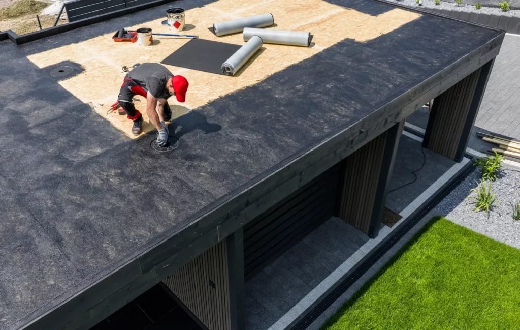 A professional roofing contractor in a red cap installing a drainage system on a flat roof with black waterproof membrane and plywood decking.