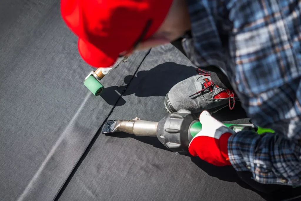 A commercial roofing specialist kneeling on a flat roof, using a professional heat welding tool and roller to join waterproof roofing membranes.