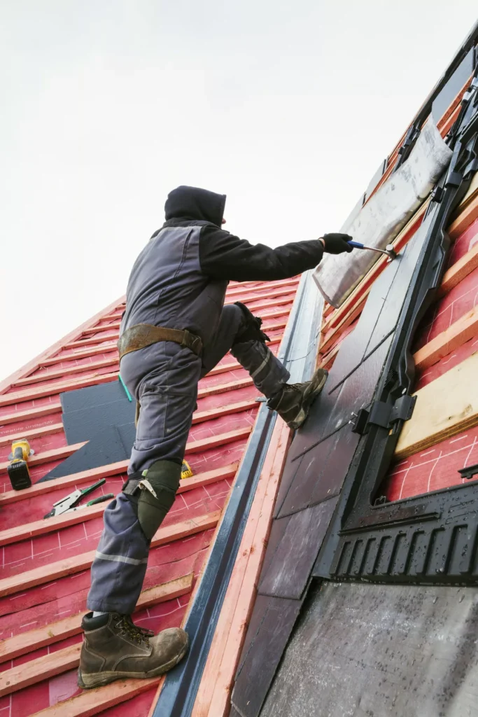 A roofing specialist in protective gear installing modern black and grey shingles over high-quality red waterproof sheathing on a residential roof.
