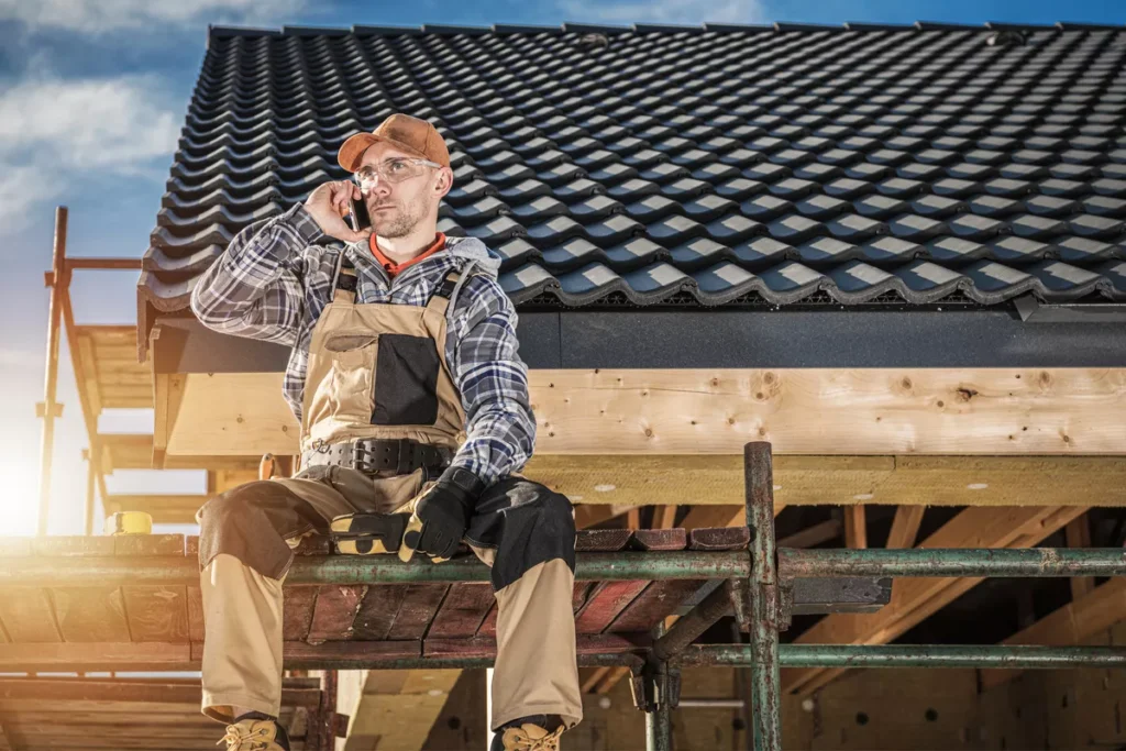 A professional roofer in safety gear and plaid shirt sitting on scaffolding while coordinating a project over the phone at a Scottsdale construction site.